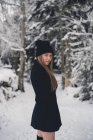 Mujer atractiva joven en sombrero divertido y abrigo negro posando en el camino del bosque y mirando a la cámara - foto de stock