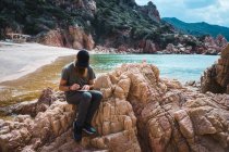 Взрослый бородатый мужчина сидит на камне и делает заметки на берегу моря. — стоковое фото