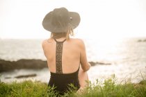 Mulher de chapéu e roupa de banho sentado na grama na costa e olhando para a vista — Fotografia de Stock