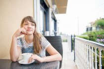 Вдумчивая молодая женщина, сидящая с кофе в кафе на открытом воздухе — стоковое фото