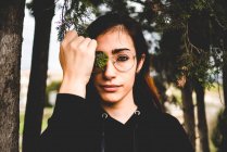 Young brunette woman in glasses covering eye with green branch in forest — Stock Photo