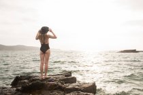 Woman in black swimsuit and hat standing on rock in sea — Stock Photo