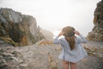 Femme en chemise et chapeau debout sur la côte rocheuse et regardant la vue — Photo de stock