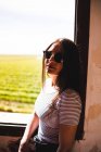 Young woman in sunglasses standing near window and looking at beautiful green field on sunny day. — Stock Photo
