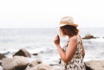 Vista posterior de la mujer tatuada en sombrero de pie sobre piedras con las manos separadas y mirando al océano - foto de stock