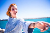 Cheerful redhead woman pulling hand and looking away while leaning on handrail at seaside. — Stock Photo