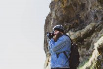 Vista lateral do homem barbudo de chapéu com mochila tirando foto por câmera da paisagem da Islândia — Fotografia de Stock