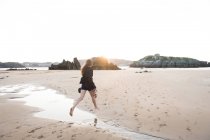 Mulher descalça segurando chapéu e calçado enquanto saltando sobre o fluxo na praia arenosa — Fotografia de Stock