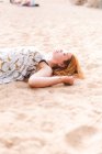 Vue latérale de la femme des cultures donnant des coups de pied de sable tout en étant couché et relaxant sur la côte sablonneuse — Photo de stock