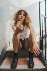 Young curly woman in brutal boots and shorts sitting on shabby stairway and showing middle fingers — Stock Photo