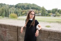 Young brunette in black dress and eyeglasses leaning on concrete wall and looking at camera — Stock Photo