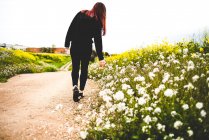 Girl in black outfit standing on remote rural road and touching flowers — Stock Photo