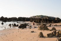 Silueta de mujer en traje de baño caminando por la playa de arena con rocas - foto de stock