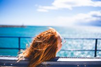 Вид сзади красивой рыжей женщины, сидящей и отдыхающей на скамейке у синего океана в солнечный день. — стоковое фото