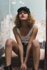 De baixo tiro da jovem mulher encaracolada em calções jeans e top tanque inclinado na cerca e olhando para cima sensualmente — Fotografia de Stock