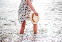 Back view of pretty woman holding hat and standing at calm ocean on beach in the bay — Stock Photo