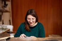 Konzentrierte Frau in grüner Bluse und Brille schnitzt Dekoration mit Instrument am Arbeitstisch in der Werkstatt — Stockfoto