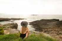 Woman in hat and swimwear sitting on grass on coast and looking at view — Stock Photo