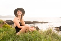 Mulher alegre em chapéu e roupa de banho sentado na grama na costa — Fotografia de Stock