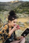 Junge hübsche Frau liest Buch und trinkt unterwegs Kaffee auf Steinen — Stockfoto