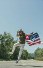 Donna felice che cammina con una bandiera americana e festeggia su una strada solitaria. Giornata speciale per festeggiare il 4 luglio — Foto stock