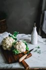Cauliflowers with green leaves lying on wooden cutting board with knife on marble table with herbs and vessel with textile material on grey room background in soft focus — Stock Photo