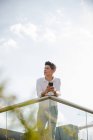 Giovane ragazzo in abito casual tenendo smartphone e guardando lontano mentre si appoggia su ringhiera su sfondo di cielo nuvoloso — Foto stock