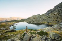 Scenic view at distance of woman standing on gray rocks on green shore of small calm lake in beautiful mountain range — Stock Photo