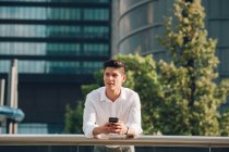 Young businessman with smartphone leaning on railing in front of modern building — Stock Photo