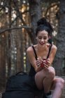 Giovane bruna sottile che tiene una tazza di metallo e siede a terra in boschi con lo zaino — Foto stock