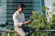 Handsome young guy in elegant outfit sitting on railing on city street and using smartphone — Stock Photo