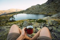 Cropped of man holding metal mug in hands while sitting on rocky coast of small lake in mountains, Spain — Stock Photo