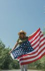 Mujer feliz caminando con una bandera americana y celebrando en un camino solitario. Día especial para celebrar el 4 de julio - foto de stock