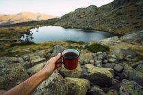 Cropped of man holding metal mug in hand while sitting on rocky coast of small lake in mountains, Spain — Stock Photo