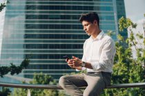 Красивый молодой парень в элегантном наряде сидит на перилах на городской улице и пользуется смартфоном — стоковое фото