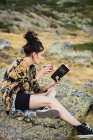 Seitenansicht einer jungen Frau, die Kaffee trinkt, während sie auf Felsen im Tal sitzt und Buch liest — Stockfoto