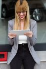 Mujer joven sonriente en gafas y estilo de negocio usando tableta con coche en el fondo - foto de stock
