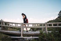 Vista laterale della donna casual con zaino rosso che cammina sul ponte pedonale in legno nella valle verde contro il cielo blu — Foto stock