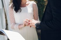 Vista de la cosecha de la novia joven en vestido de novia y el novio en traje negro intercambio de anillos en la ceremonia en el jardín - foto de stock