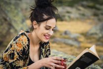 Junge hübsche lächelnde Frau liest Buch auf Steinen auf Reisen — Stockfoto
