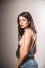 Сексуальна дівчина в джинсовому комбінезоні над голим тілом позує на сірому фоні — стокове фото