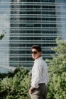 Young businessman in sunglasses standing in front of modern building and looking over shoulder — Stock Photo