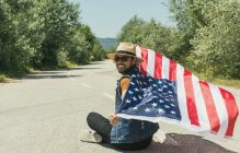 Homem com bandeira americana sentado na estrada — Fotografia de Stock