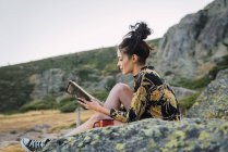 Junge brünette Frau liest Buch auf Steinen auf Reisen — Stockfoto