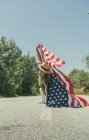 Femme heureuse marchant avec un drapeau américain et célébrant sur une route solitaire. Journée spéciale pour célébrer le 4 juillet — Photo de stock