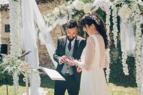 Young beautiful bride in white dress and handsome groom in black costume exchanging rings on background of wedding decorations — Stock Photo