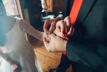 Crop view of man in black costume helping young bride in wedding gown put on bracelet on hand on background of room - foto de stock