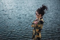 Side view of woman in colorful shirt embracing herself and looking away standing on blurred background of blue water in ripples — Stock Photo