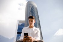 Молодий хлопець у повсякденному вбранні, використовуючи смартфон на тлі хмарного неба та сучасного хмарочоса — стокове фото