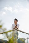 Junger Kerl in lässigem Outfit hält Smartphone in der Hand und schaut weg, während er sich an Geländer vor wolkenverhangenem Himmel lehnt — Stockfoto
